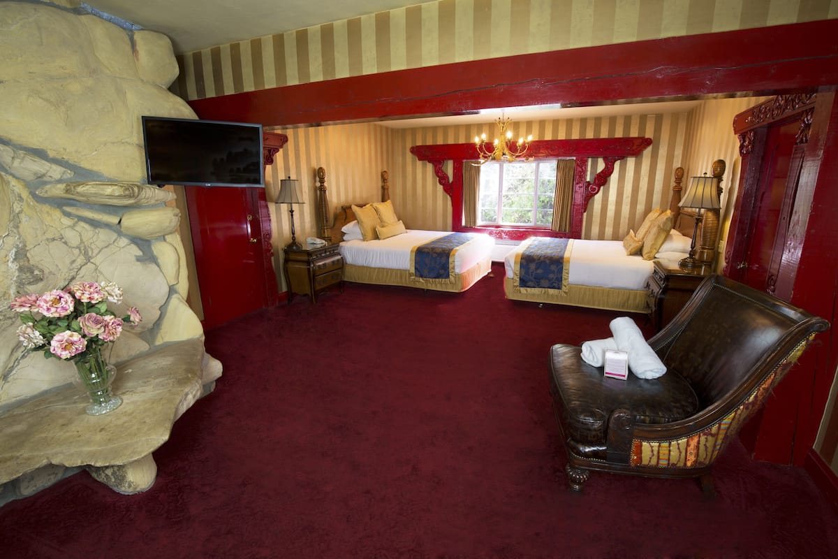 Ein Hotelzimmer mit zwei Betten, einem Fernseher, einer Steinwand, einem roten Teppich und einem Sessel mit Handtüchern.
