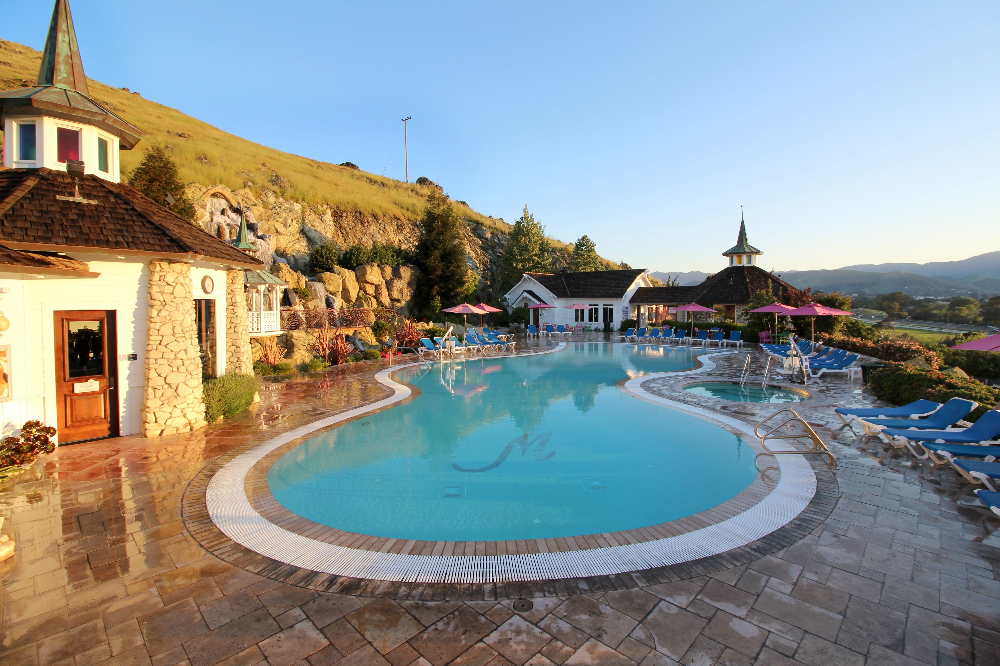 A swimming pool with lounge chairs and umbrellas, surrounded by stone buildings and a view of hills.