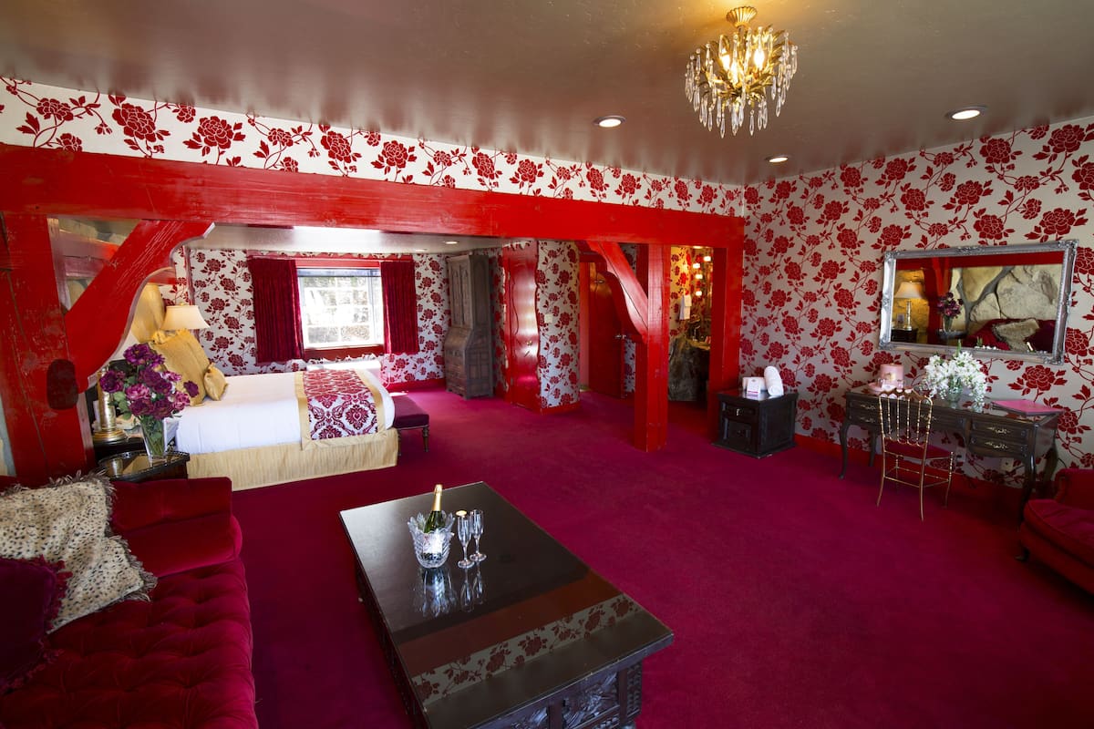 Ein Zimmer mit rot-weißer floraler Tapete, einem Chandelier, einem Bett mit rotem Baldachin, einem roten Sofa und einem Schreibtisch mit Spiegel.