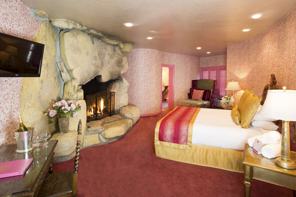 Ein Hotelzimmer mit Kamin aus Stein, einem Bett mit roter und goldener Bettdecke, einem Nachttisch mit Lampe und einem an der Wand befestigten Fernseher.
