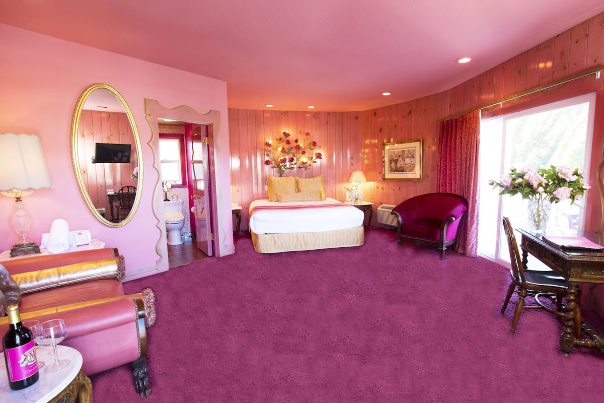 Ein hotelzimmer in Rosa gehalten mit einem großen Bett, einem Schminktisch mit Spiegel, einer Sitzecke mit einem Sessel und einem kleinen Tisch mit Flasche und Glas.