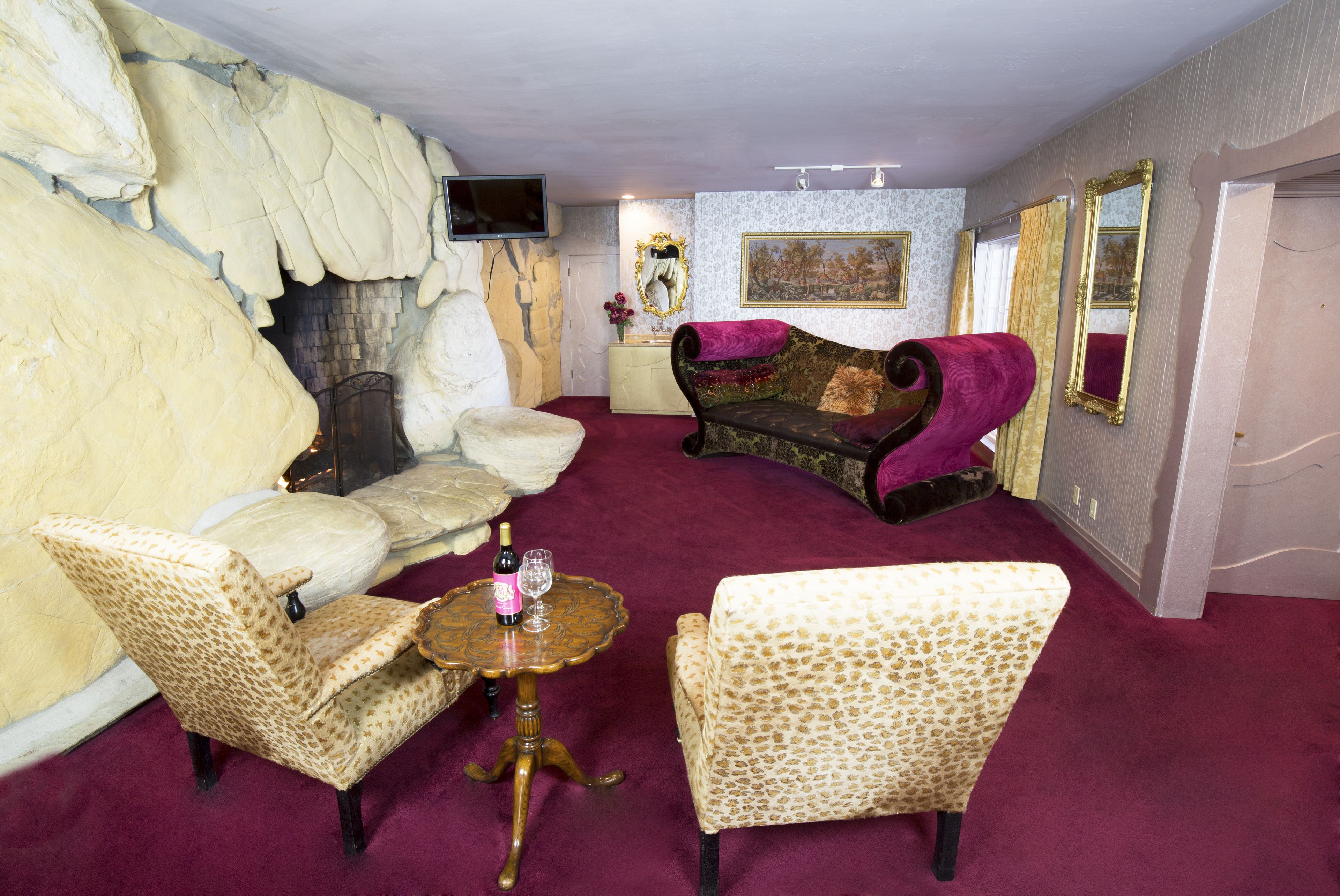 Une pièce avec un mur en pierre, une cheminée, un canapé, deux fauteuils, une petite table avec une bouteille et un verre, et un téléviseur accroché au mur.