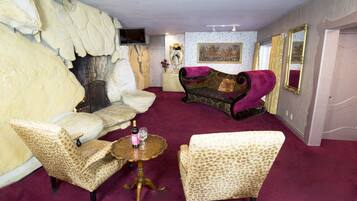 Une pièce avec un mur en pierre, une cheminée, un canapé, deux fauteuils, une petite table avec une bouteille et un verre, et un téléviseur accroché au mur.