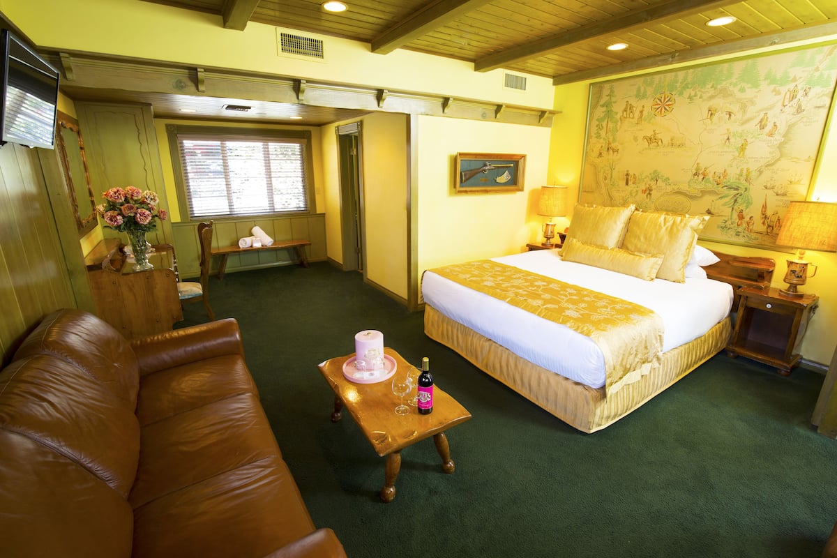 Ein Hotelzimmer mit einem großen Bett, einem Couchtisch aus Holz mit einer Flasche und Gläsern, einem braunen Ledersofa, einem kleinen Tisch mit Blumen und einer Karte an der Wand.