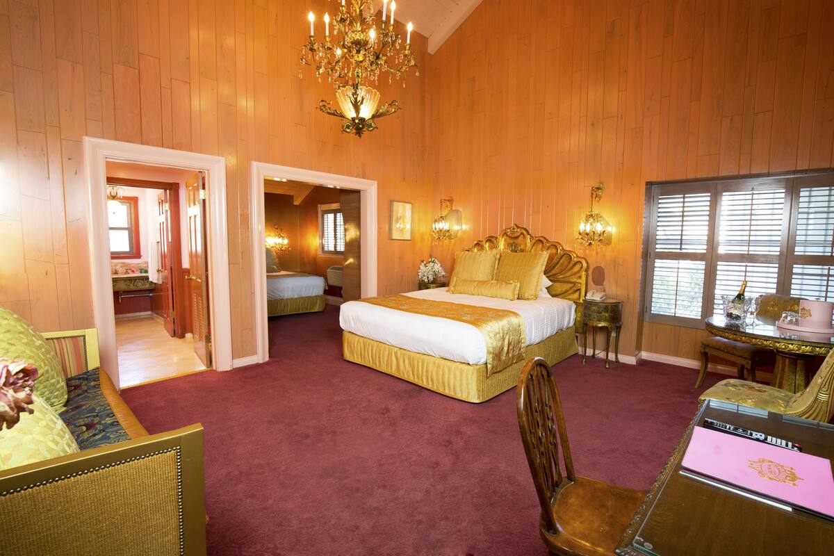 Ein geräumiges Schlafzimmer mit einem großen Bett, einem Schreibtisch und einem Sessel. Es gibt eine Chandelier, ein Fenster mit Jalousien und eine Tür zu einem anderen Raum.