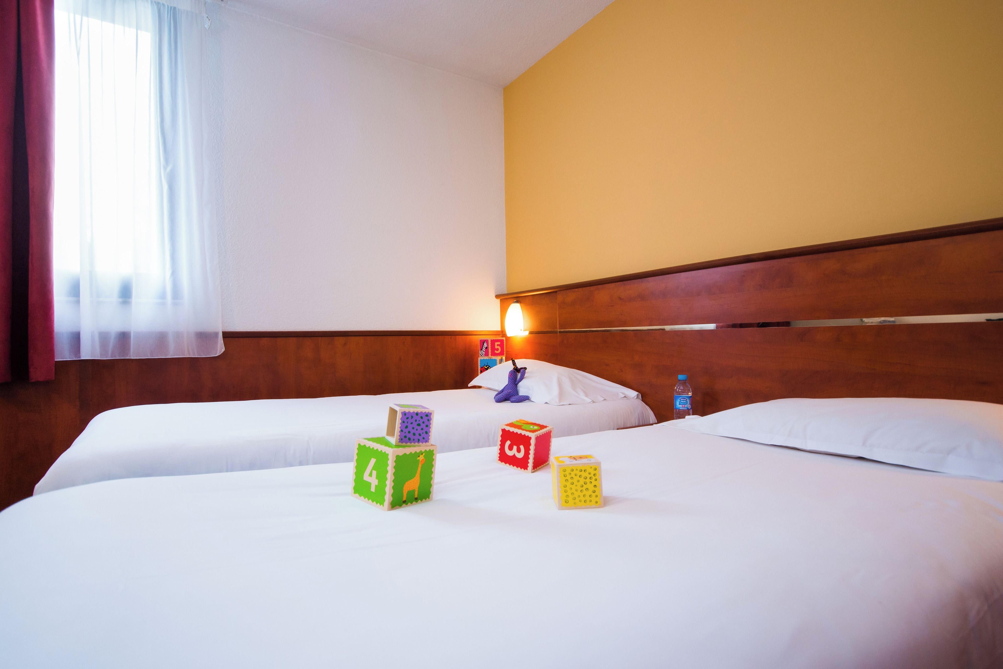 Habitación de hotel con dos camas, cabecero de madera, ventana con cortinas y lámpara fijada a la pared.