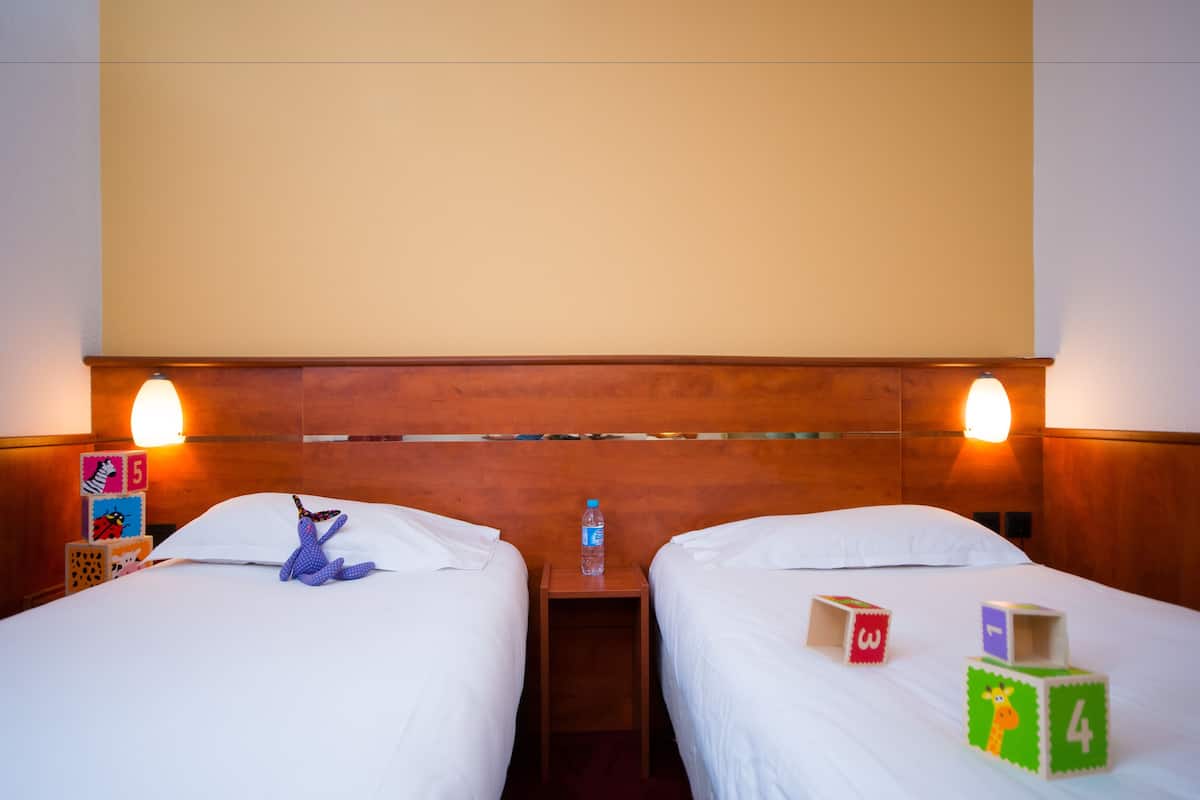 Une chambre d’hôtel avec deux lits, une tête de lit en bois, des tables de chevet avec des lampes et des jeux de construction pour enfants sur les lits.