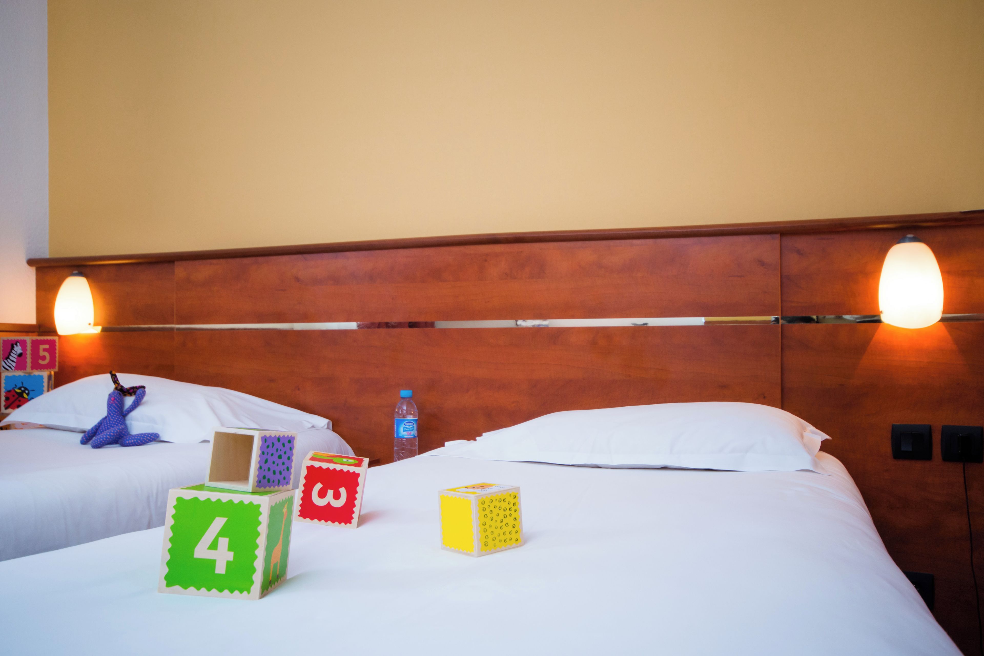 A hotel room with two beds, wooden headboard, wall-mounted lights, and children's blocks on the beds.