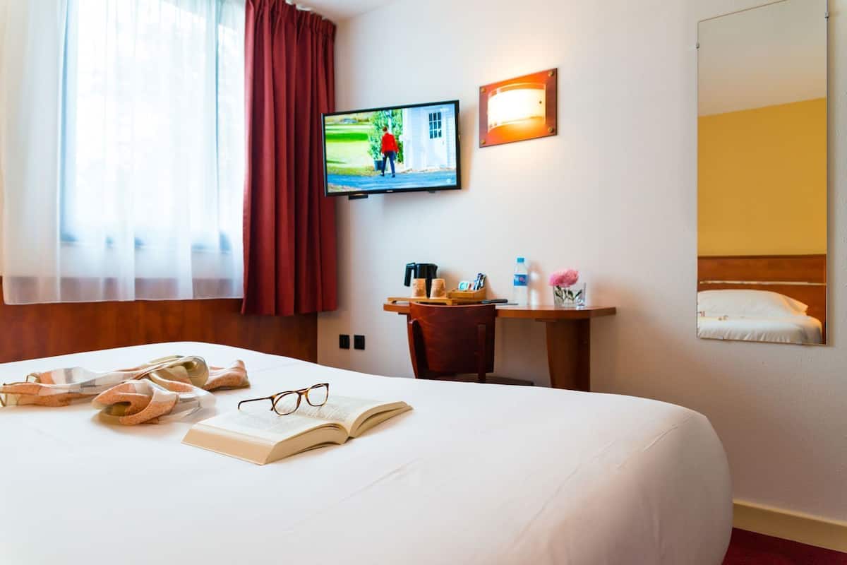 Une chambre d’hôtel équipée d’un lit, d’une télévision, d’un bureau avec une chaise, d’un miroir et d’une fenêtre avec des rideaux.
