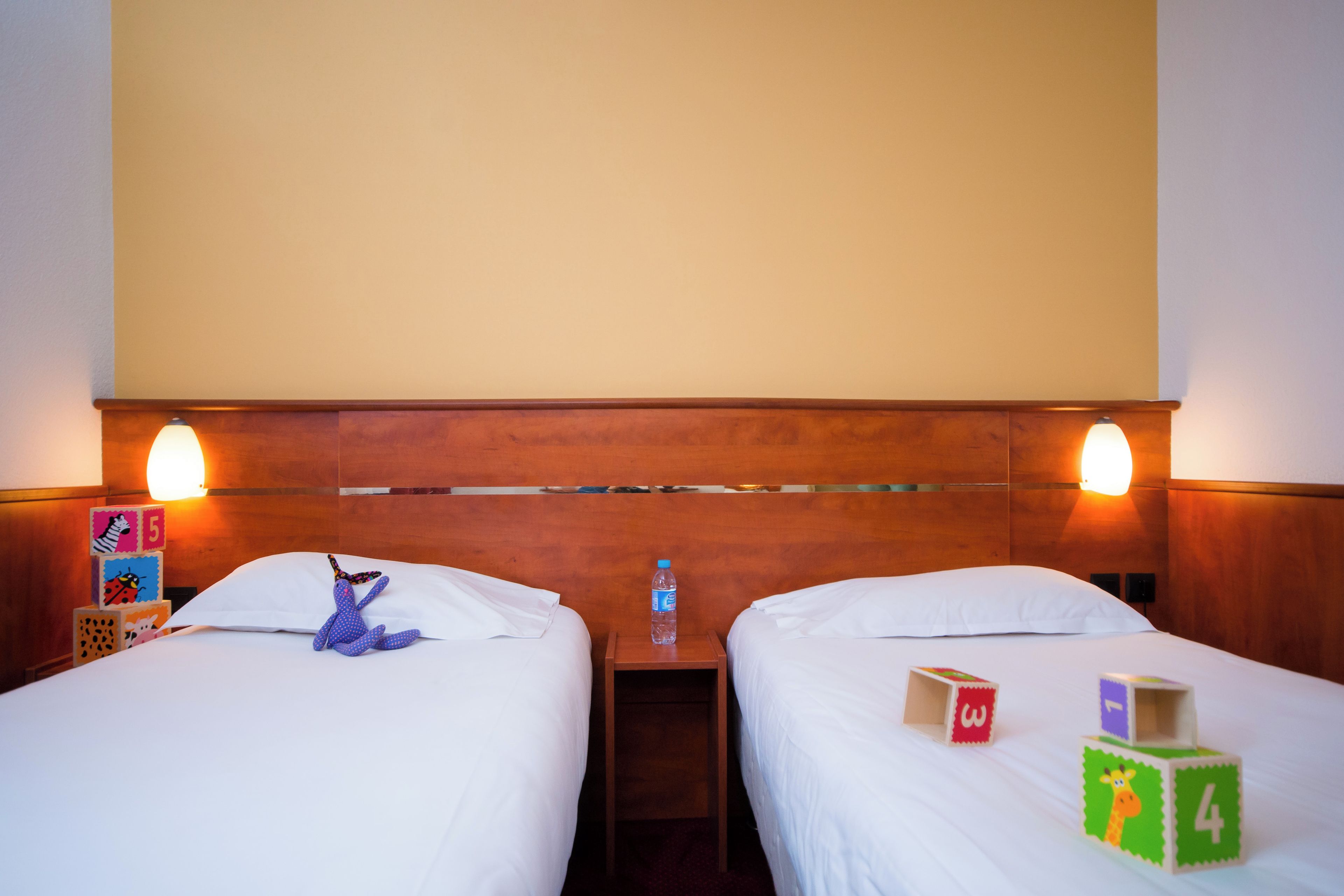 A hotel room with two beds, a wooden headboard, bedside lamps, and children's blocks on the nightstand.