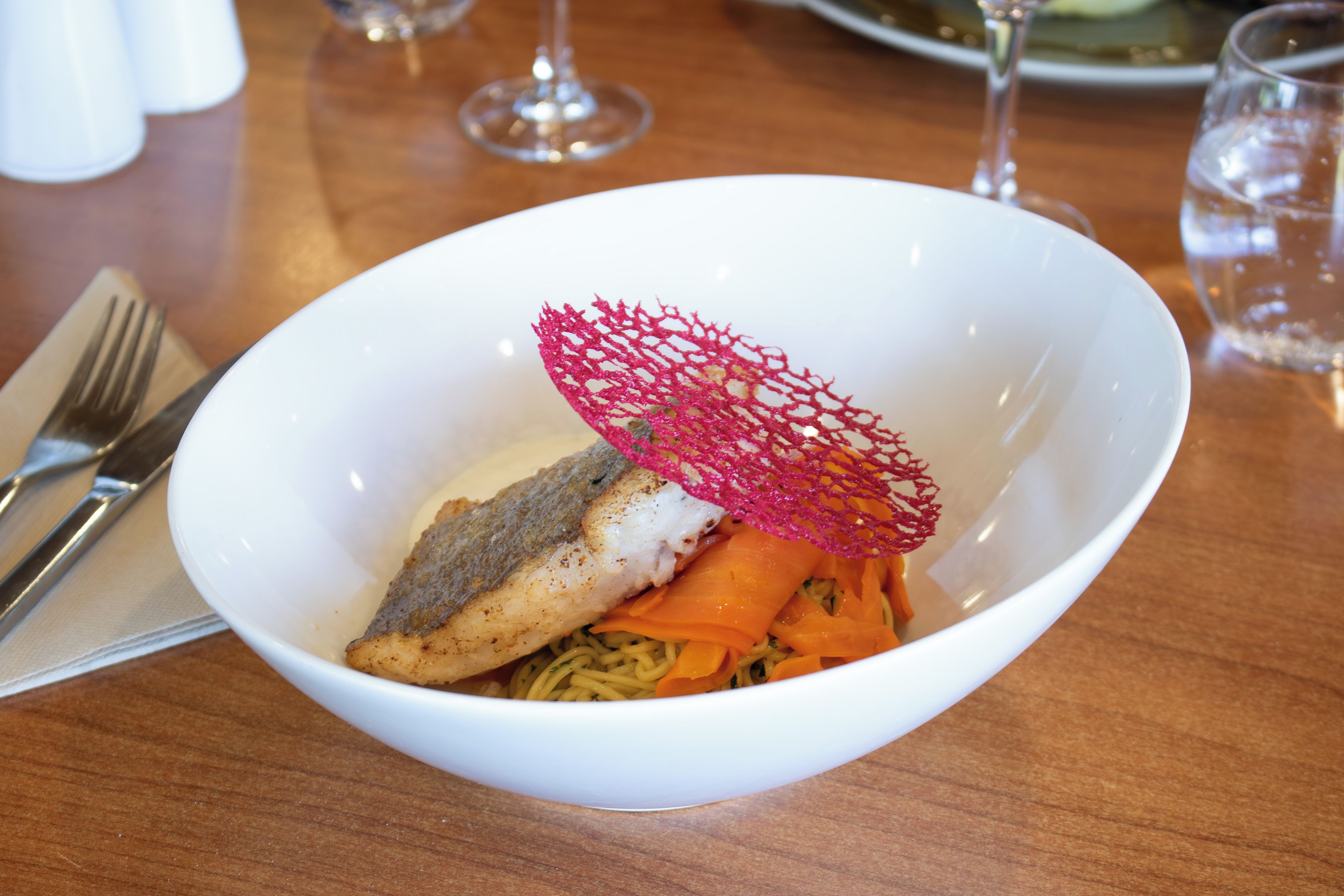 Uma tigela branca com peixe, macarrão e cenouras, decorada com uma rede vermelha.