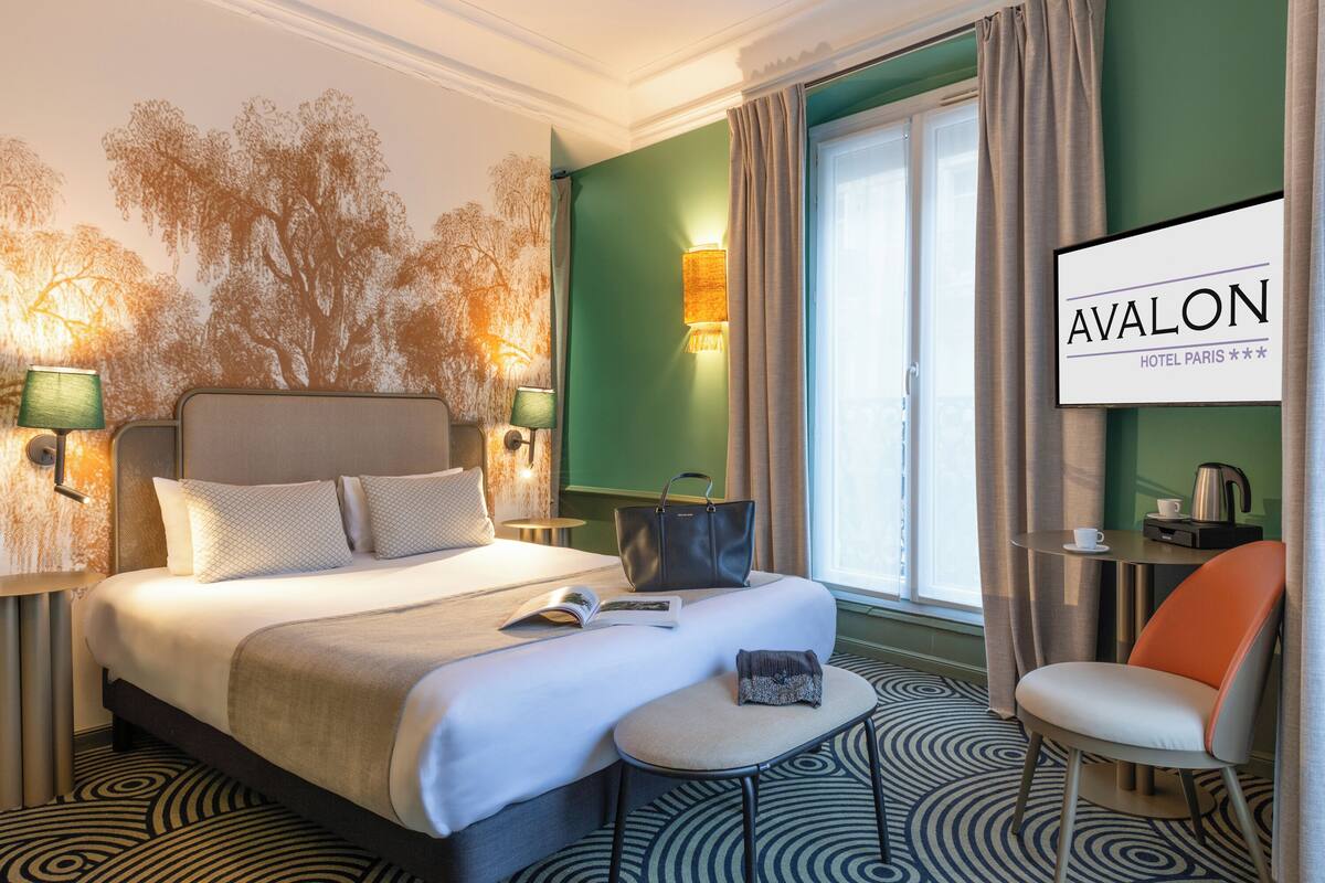 Ein Hotelzimmer mit einem großen Bett, einem Lesesessel, einem kleinen Tisch und Blick auf den Eiffelturm.