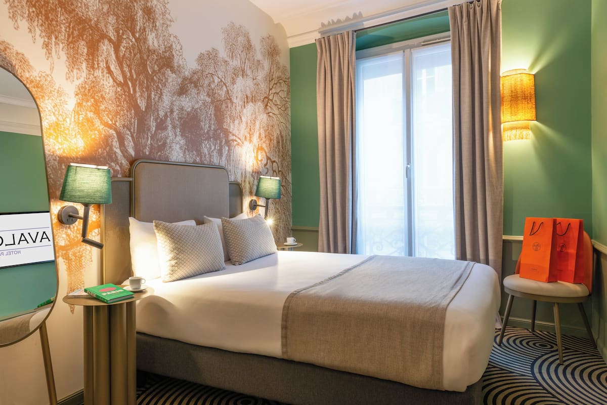Ein Hotelzimmer mit einem großen Bett, zwei Nachttischen mit Lampen, Blick ins Freie und einer Wand mit einem Baum-Wandbild.