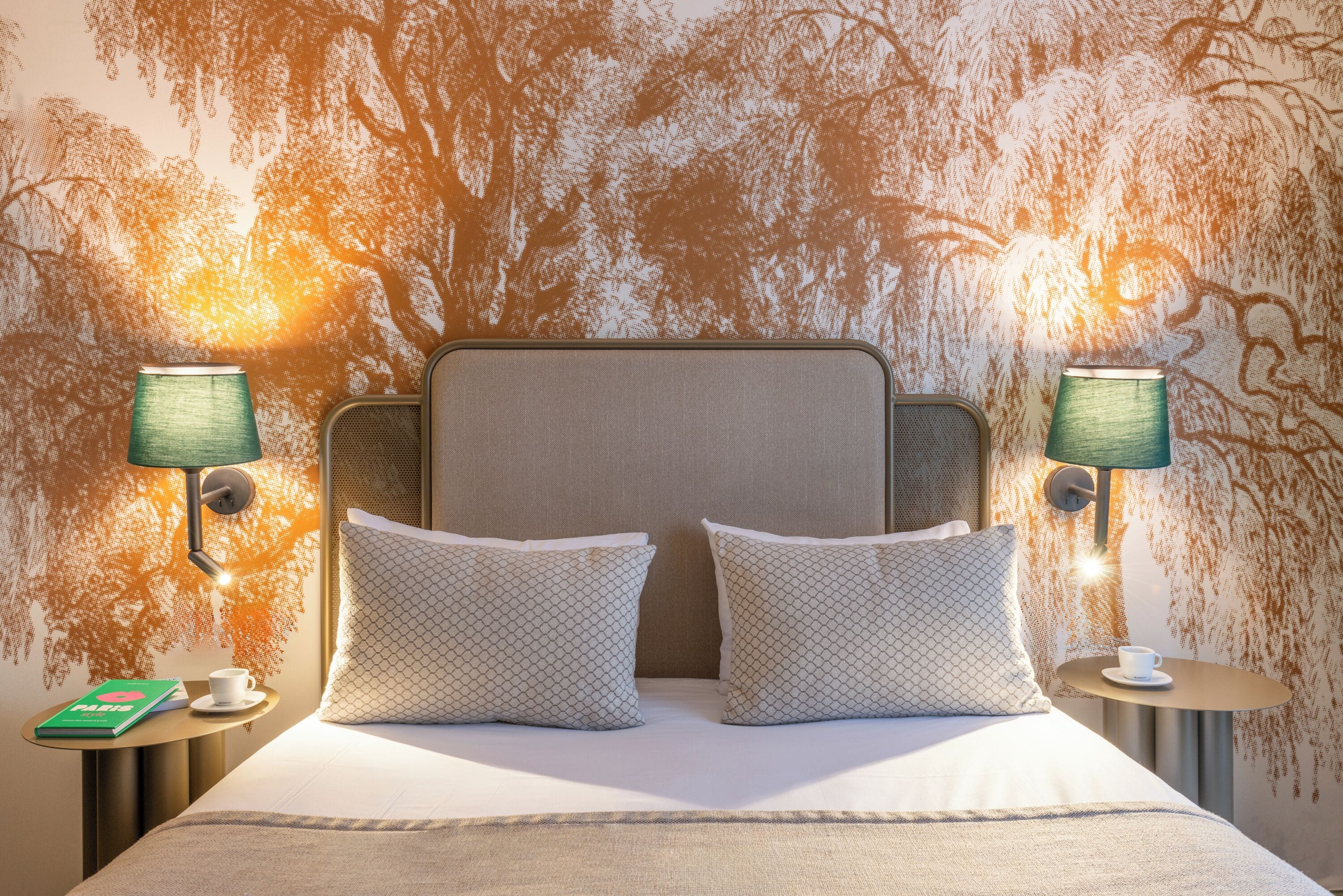 A neatly made bed with two pillows, two bedside lamps, and a small table with a cup and book.