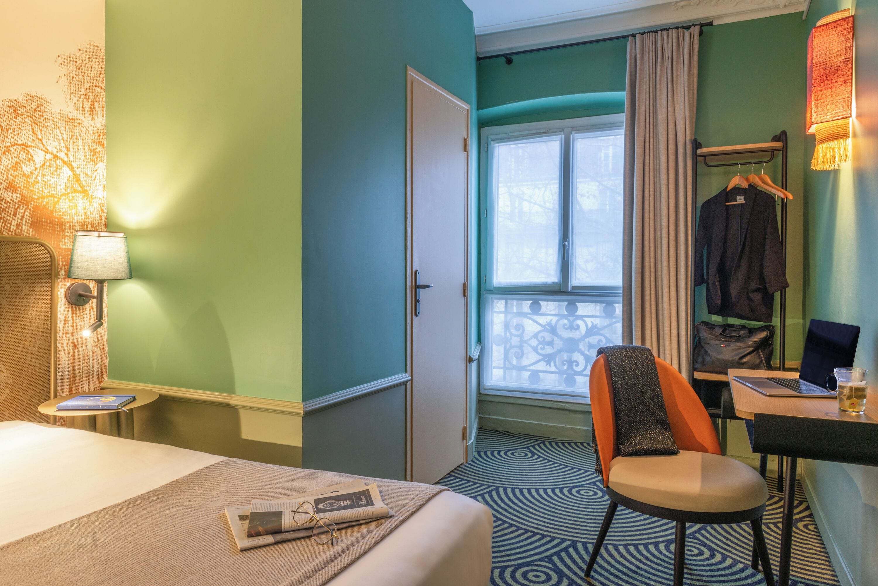 Et hotelværelse med en seng, et skrivebord med en bærbar computer, en stol og et vindue med gardiner.