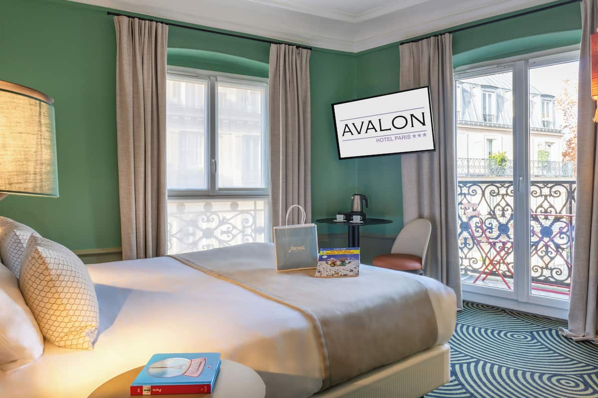 Ein Hotelzimmer mit Bett, Nachttisch mit Lampe und Blick auf den Balkon. Ein Schild mit der Aufschrift 'AVALON HOTEL PARIS'.