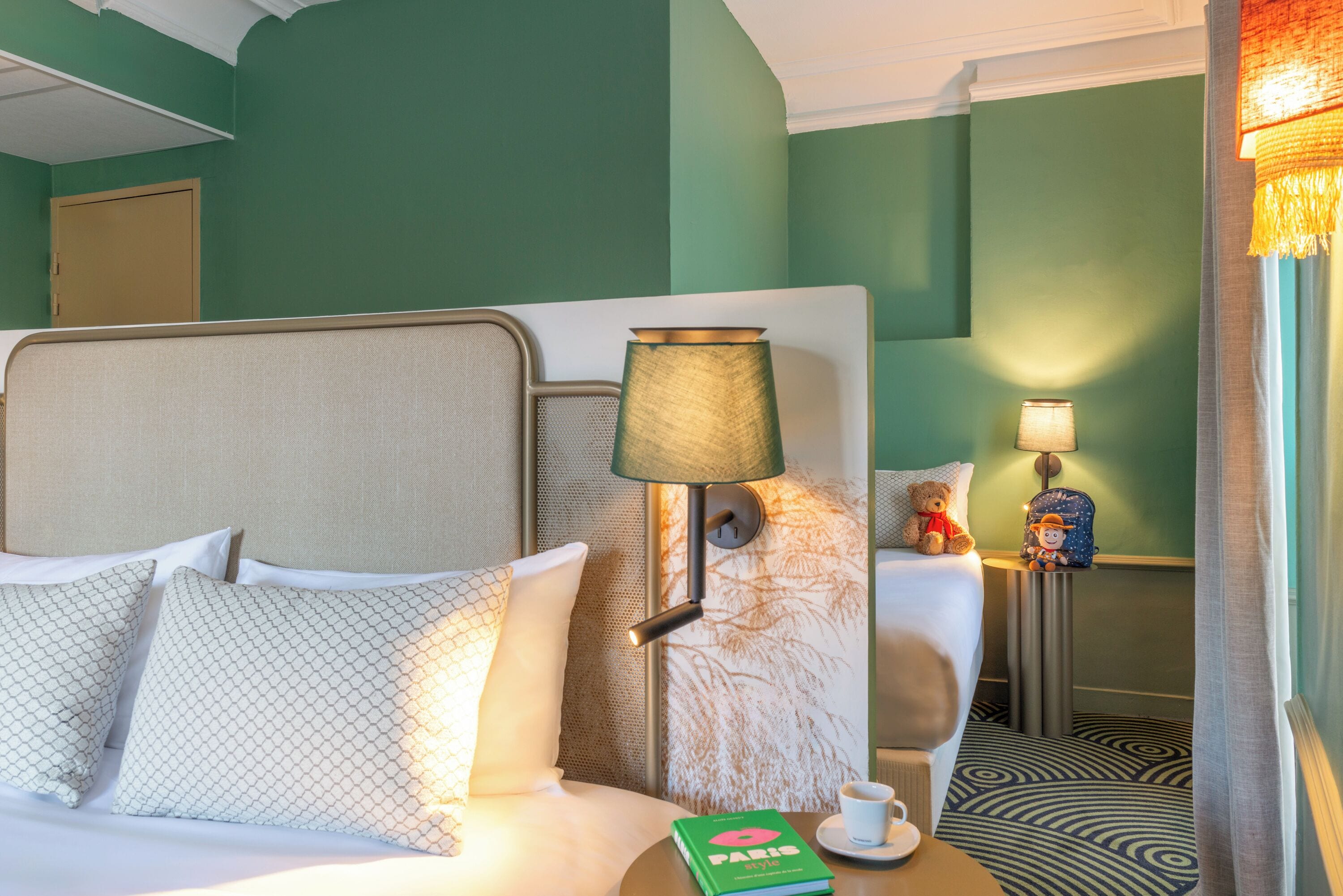 A hotel room with a bed, a nightstand, a lamp, a teddy bear, a book, and a cup.