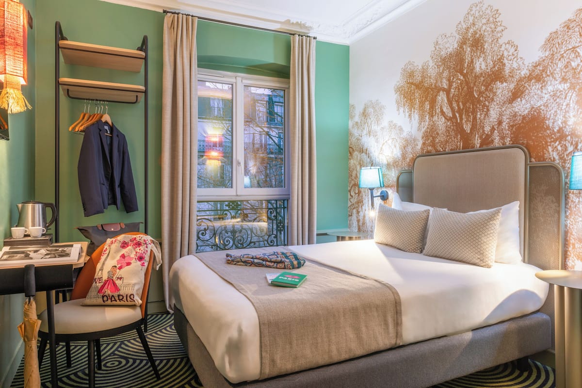 Ein Hotelzimmer mit Bett, Schreibtisch, Stuhl, einem Fenster mit Vorhängen und einer Wand mit einem Baum-Mural.