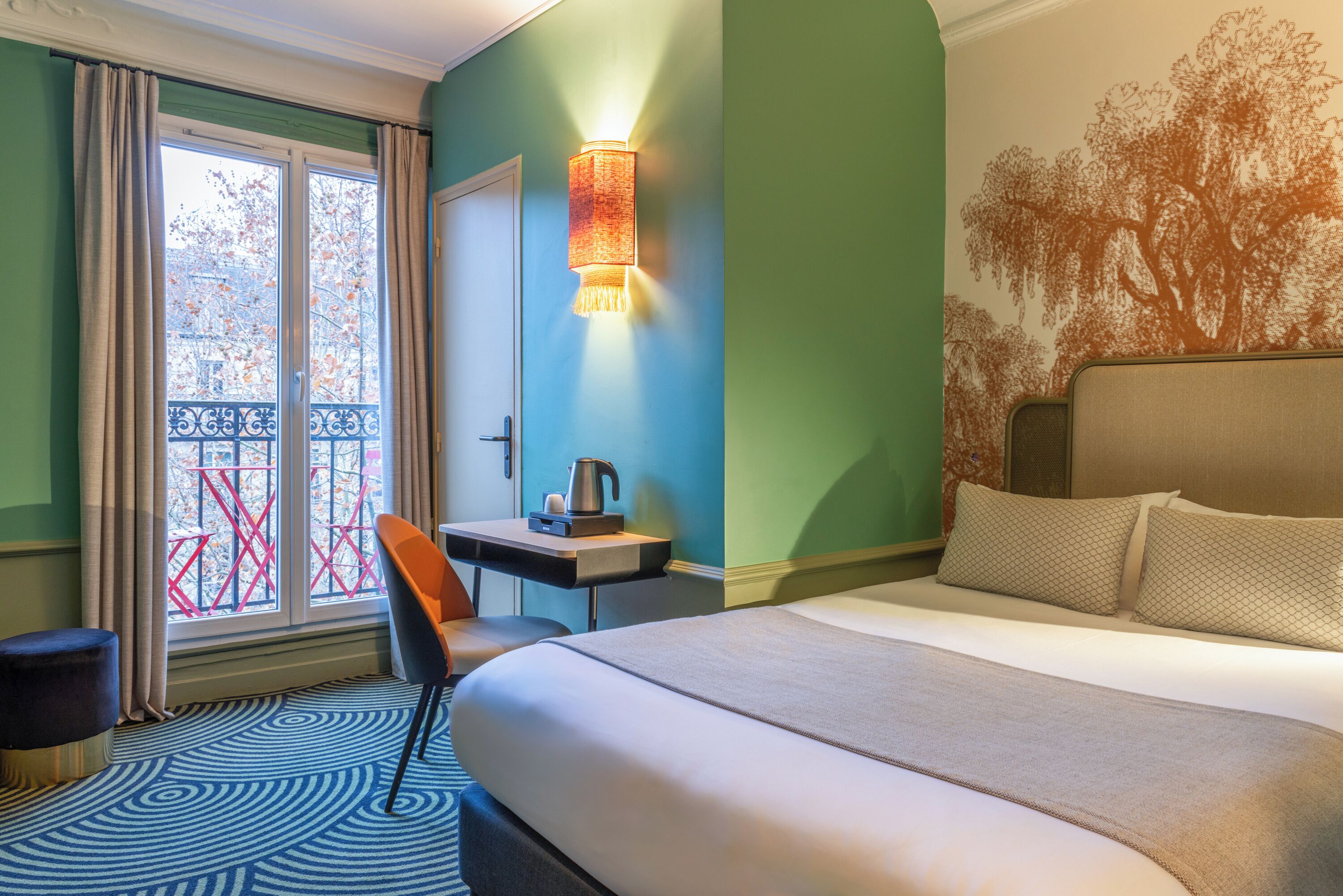 A hotel room with a bed, a desk with a kettle, a chair, and a view of a balcony with red railings.