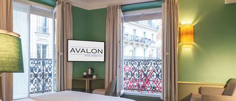 A hotel room with a bed, a chair, a small table with a lamp, and a view of the city through large windows.