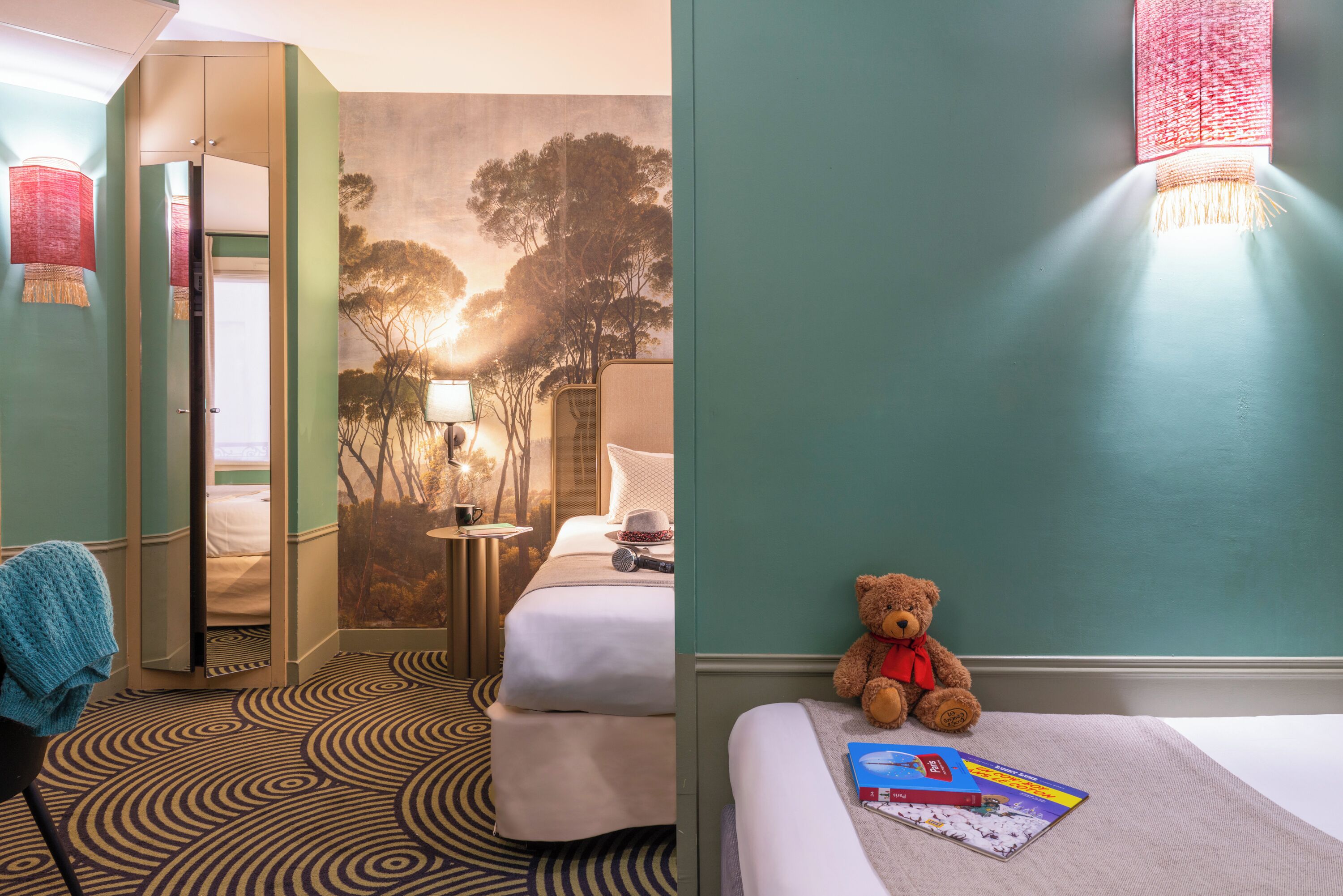 A hotel room with a bed, a chair, a teddy bear, and a wall with a tree mural.