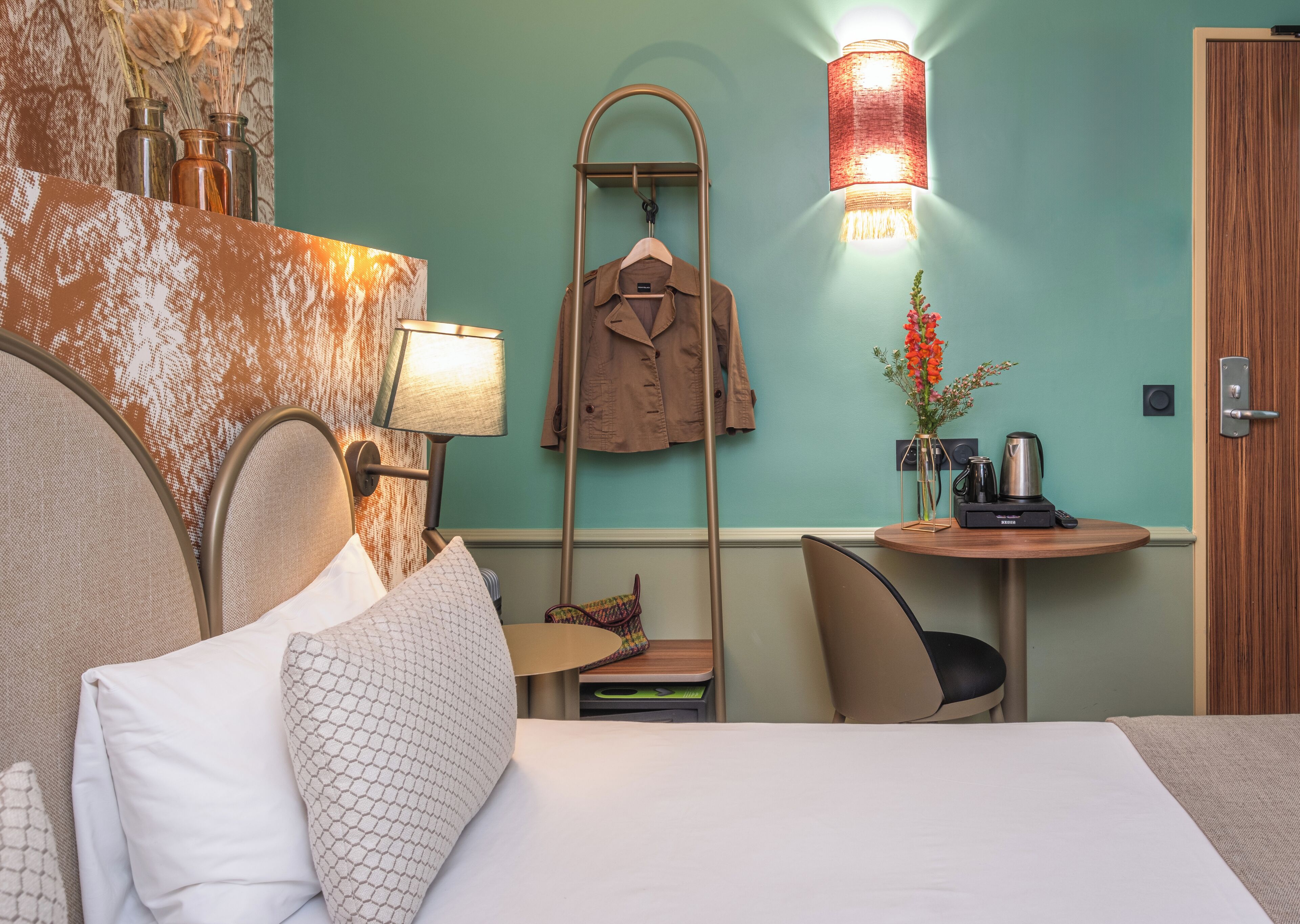 A hotel room with a bed, a desk with a lamp, a coat rack, and a wall-mounted light fixture.