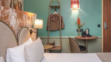 A hotel room with a bed, a desk with a lamp, a coat rack, and a wall-mounted light fixture.