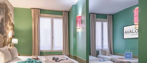 A hotel room with a large bed, a smaller bed, a chair, a desk with a laptop, and a wall-mounted lamp.