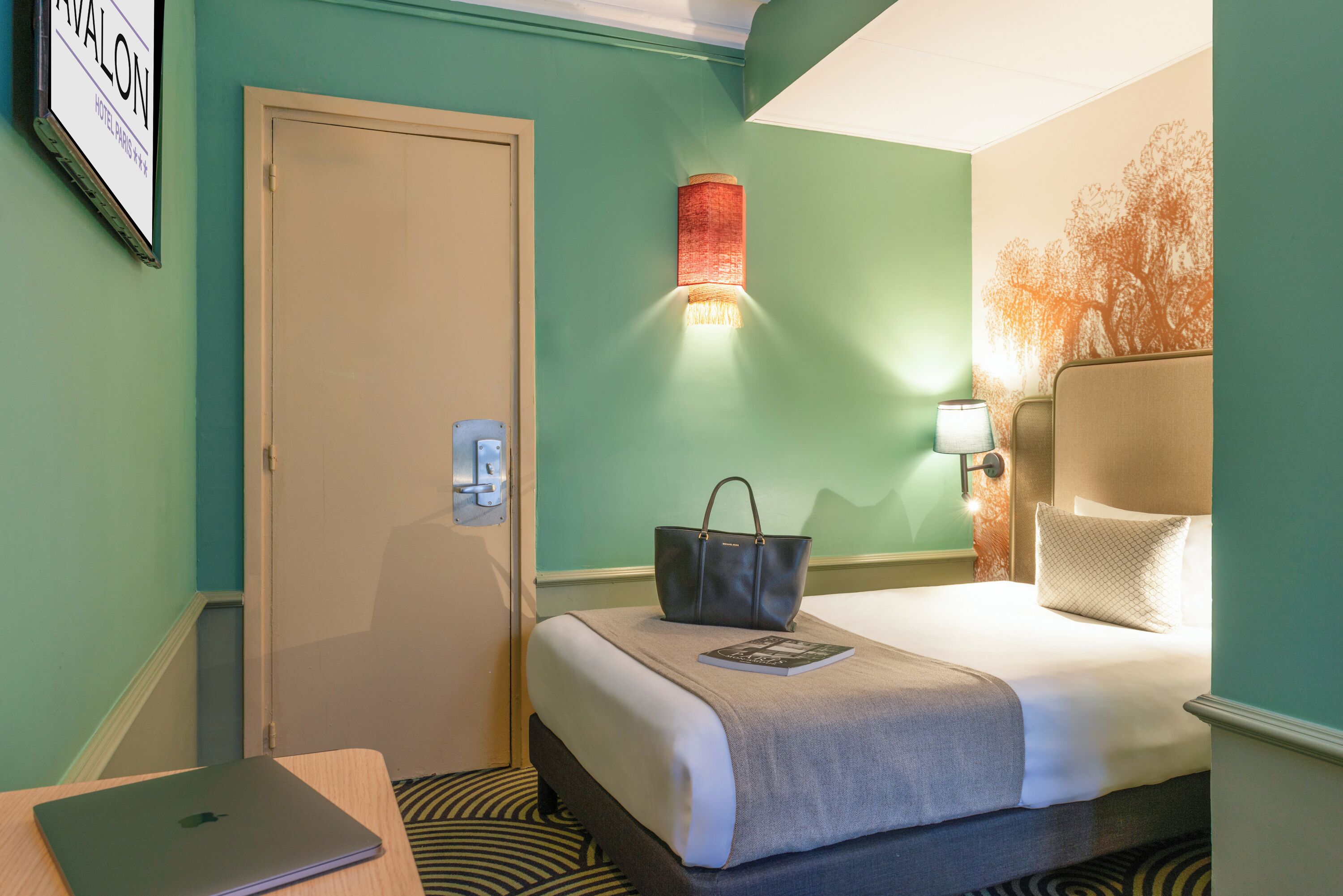 Quarto de hotel com cama, secretária com laptop, luminária na parede e porta.
