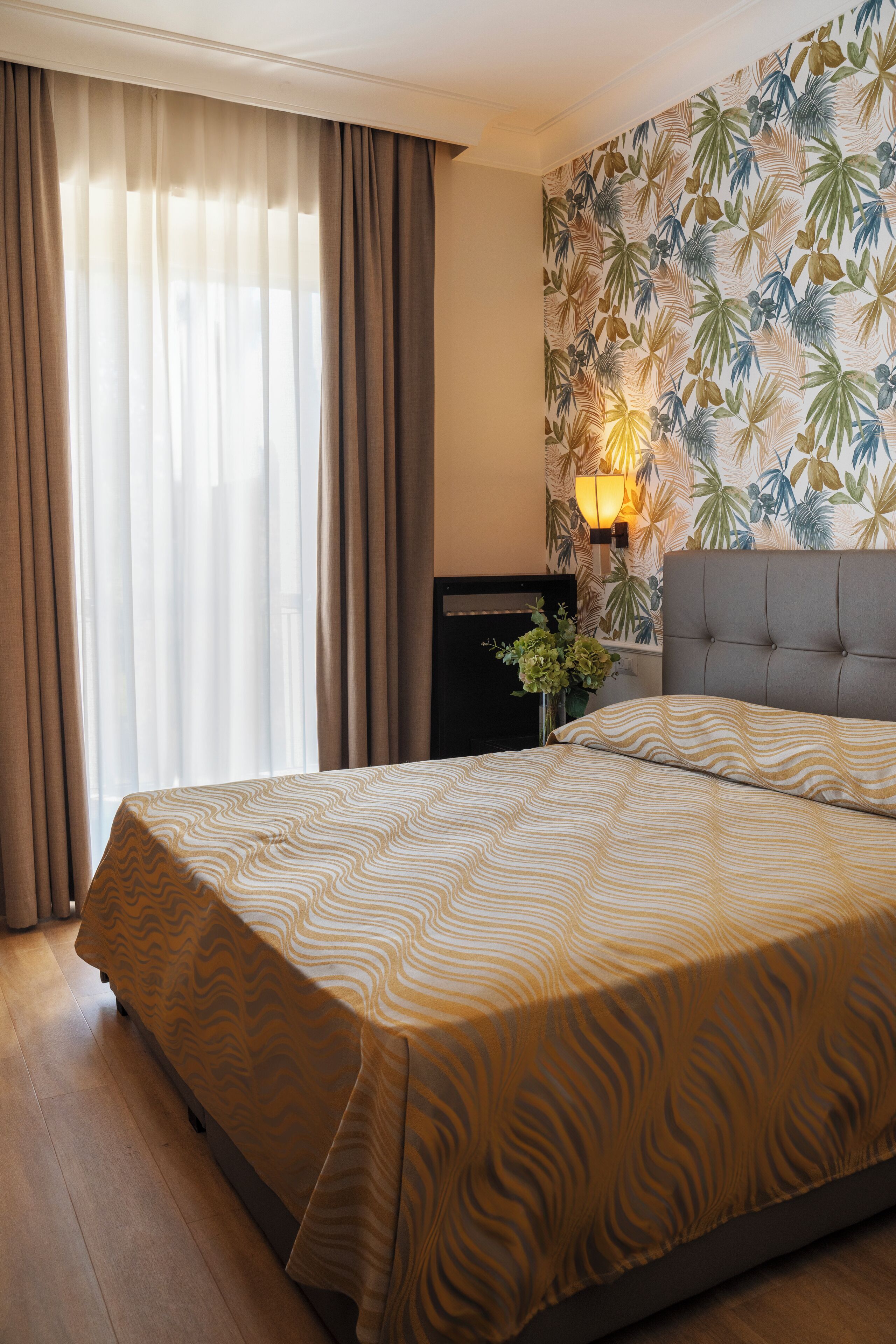 Habitación de hotel con una cama grande, cabecera, pared con diseño de hojas tropicales, mesita de noche con lámpara y ventana con cortinas.