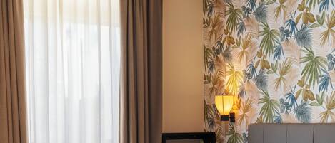 Habitación de hotel con una cama grande, cabecera, pared con diseño de hojas tropicales, mesita de noche con lámpara y ventana con cortinas.