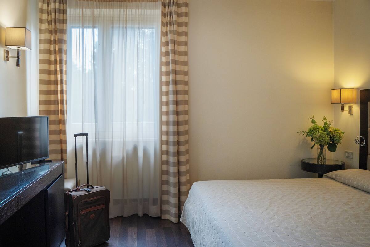 A hotel room with a bed, a TV, a suitcase, a lamp, and a vase with flowers.