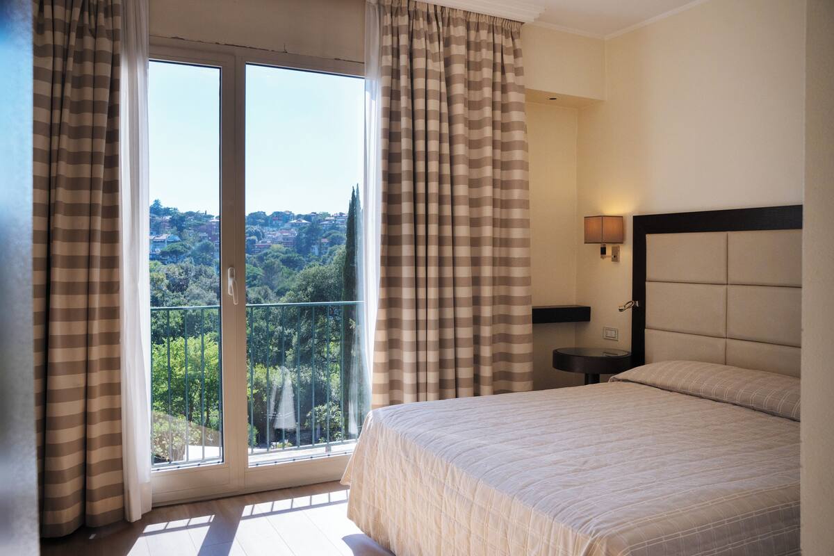 A hotel room with a large bed, a nightstand, a lamp, and a view of a balcony with curtains.