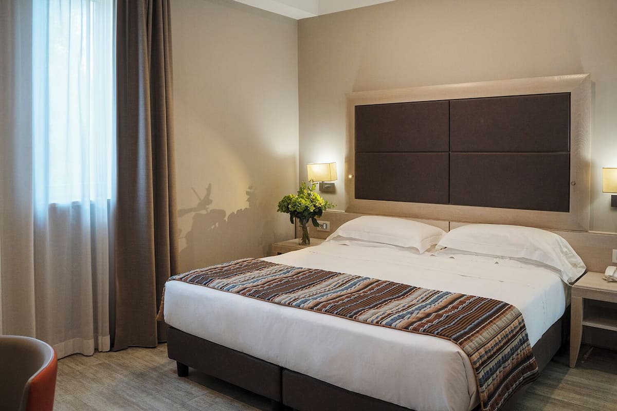 A hotel room with a large bed, a nightstand with a lamp, and a window with curtains.