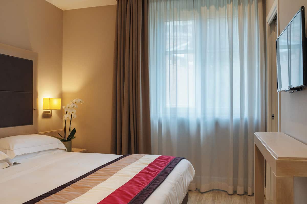 A hotel room with a bed, a nightstand with a lamp and flowers, a TV, and a window with curtains.