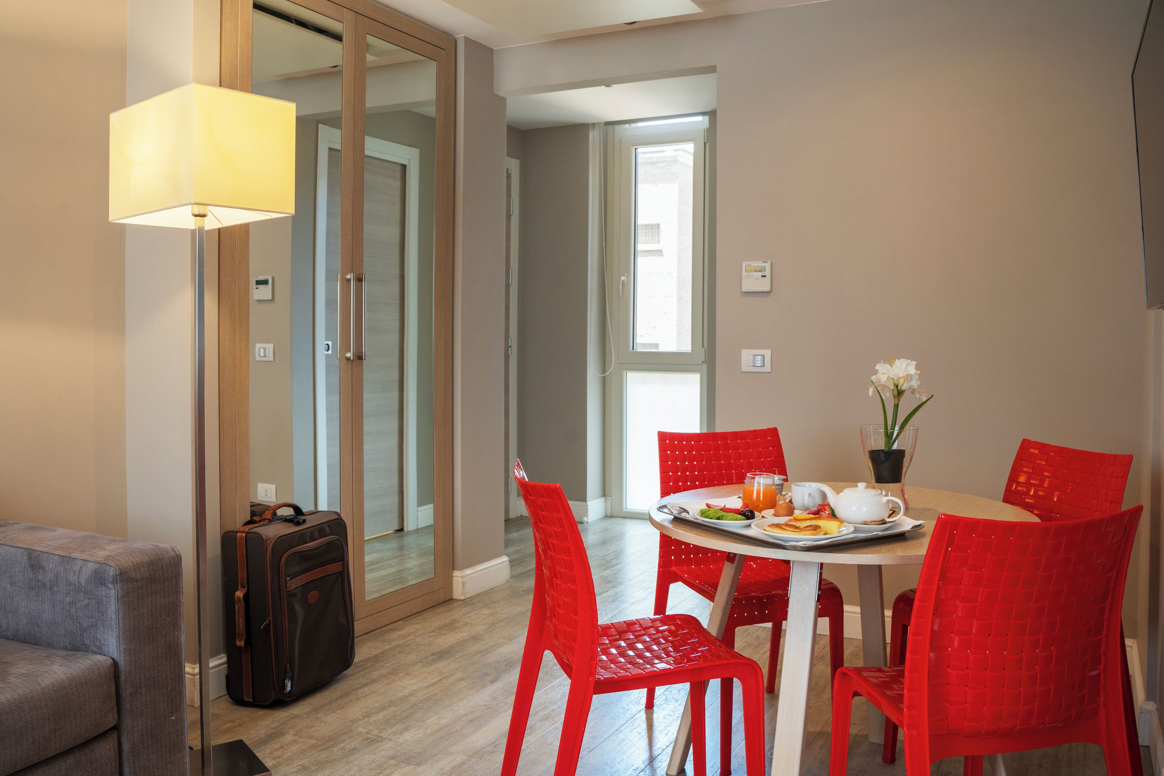 Un coin repas avec une table dressée pour deux personnes, des chaises rouges, une lampe et une valise.