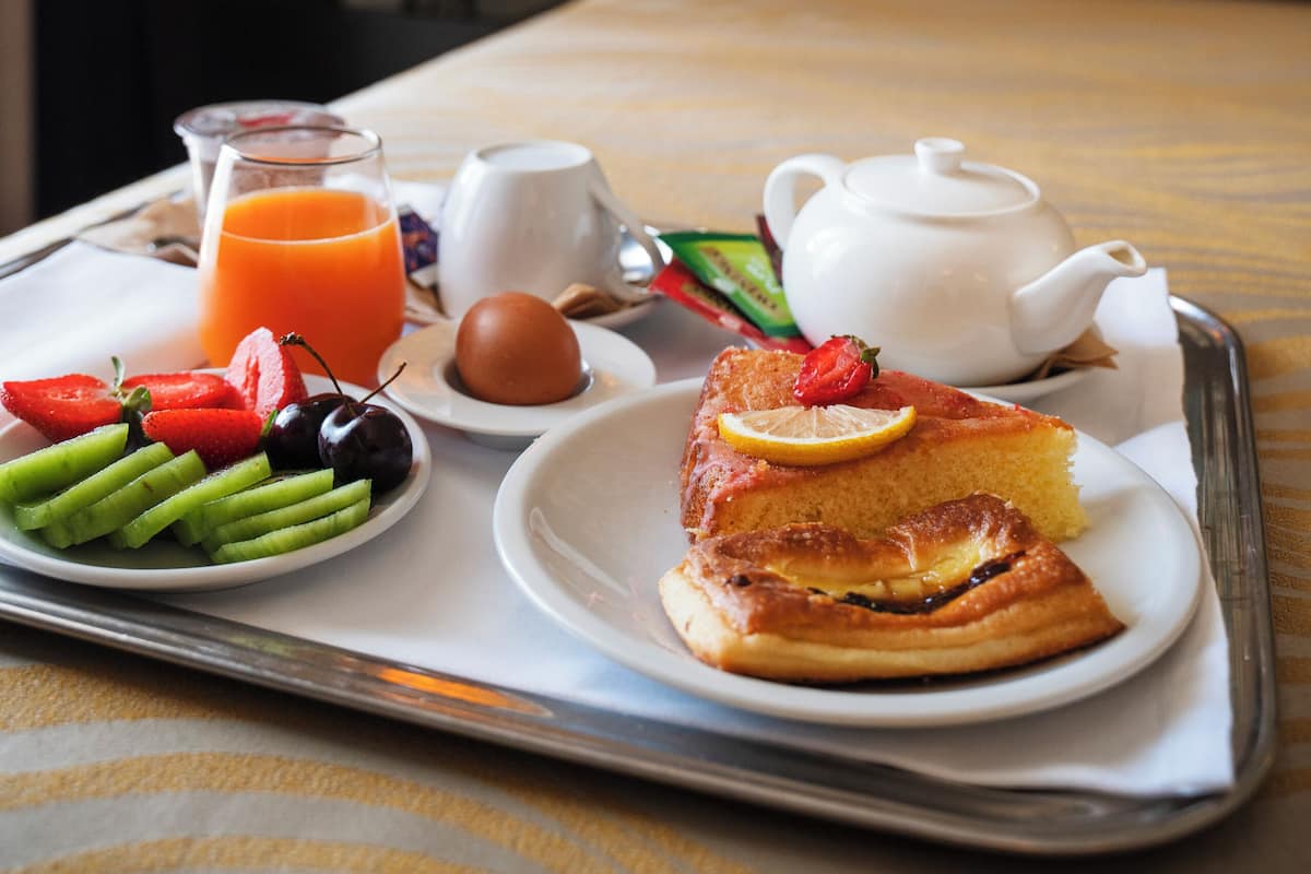 A breakfast tray with toast, fruit, eggs, and juice on a bed with a headboard and a wall with a floral pattern.