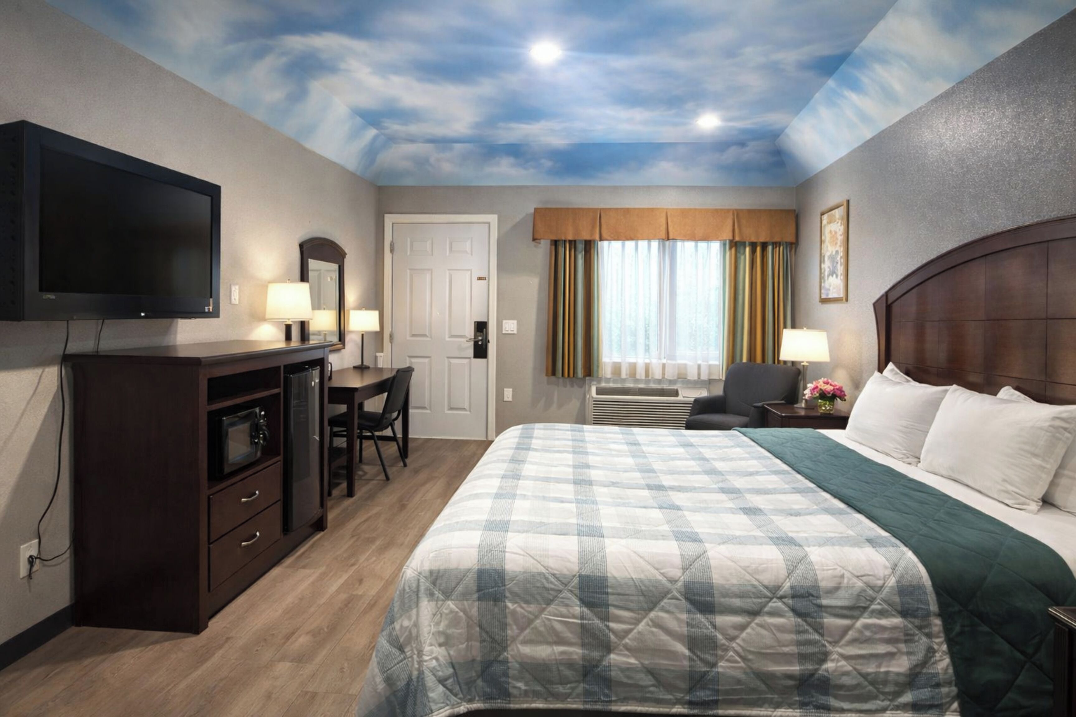 A hotel room with a large bed, a desk with a chair, a television, and a ceiling with a cloud design.