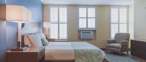 A hotel room with a bed, bedside lamps, a chair, a desk, and an air conditioning unit.