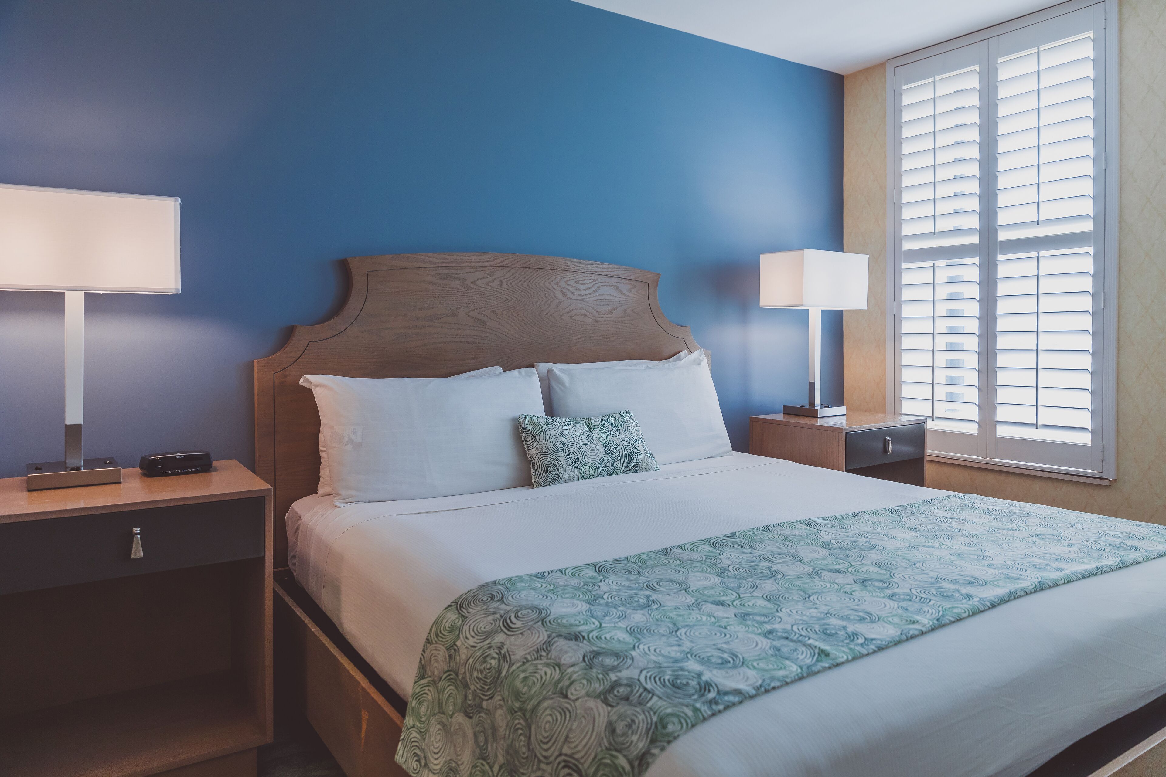 A neatly made bed with a patterned blanket, two bedside lamps, and a window with blinds.