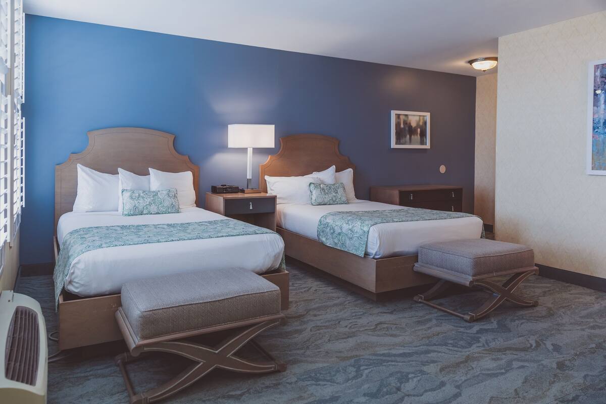 A hotel room with two beds, a bedside table with a lamp, a small cabinet, and a carpeted floor.