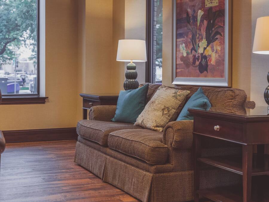 A cozy living room with a brown sofa, teal cushions, two side tables with lamps, and a large floral painting on the wall.