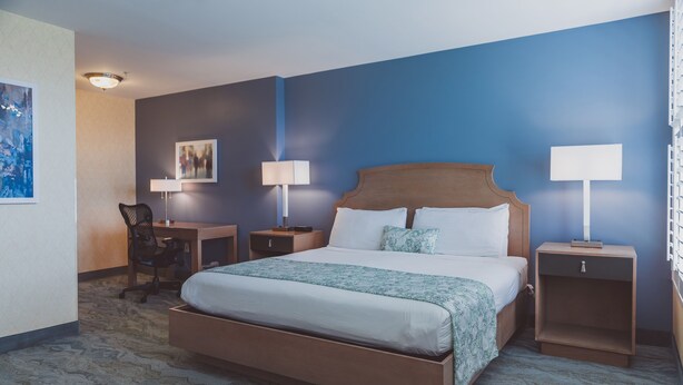 Una habitación de hotel con una cama grande, dos mesitas de noche con lámparas, un escritorio con una silla y obras de arte en las paredes.