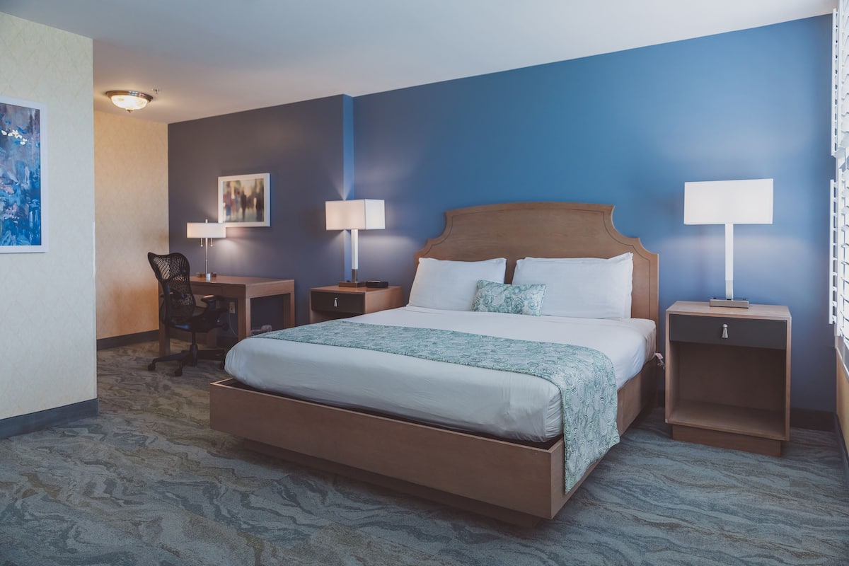 A hotel room with a large bed, two bedside tables with lamps, a desk with a chair, and artwork on the walls.
