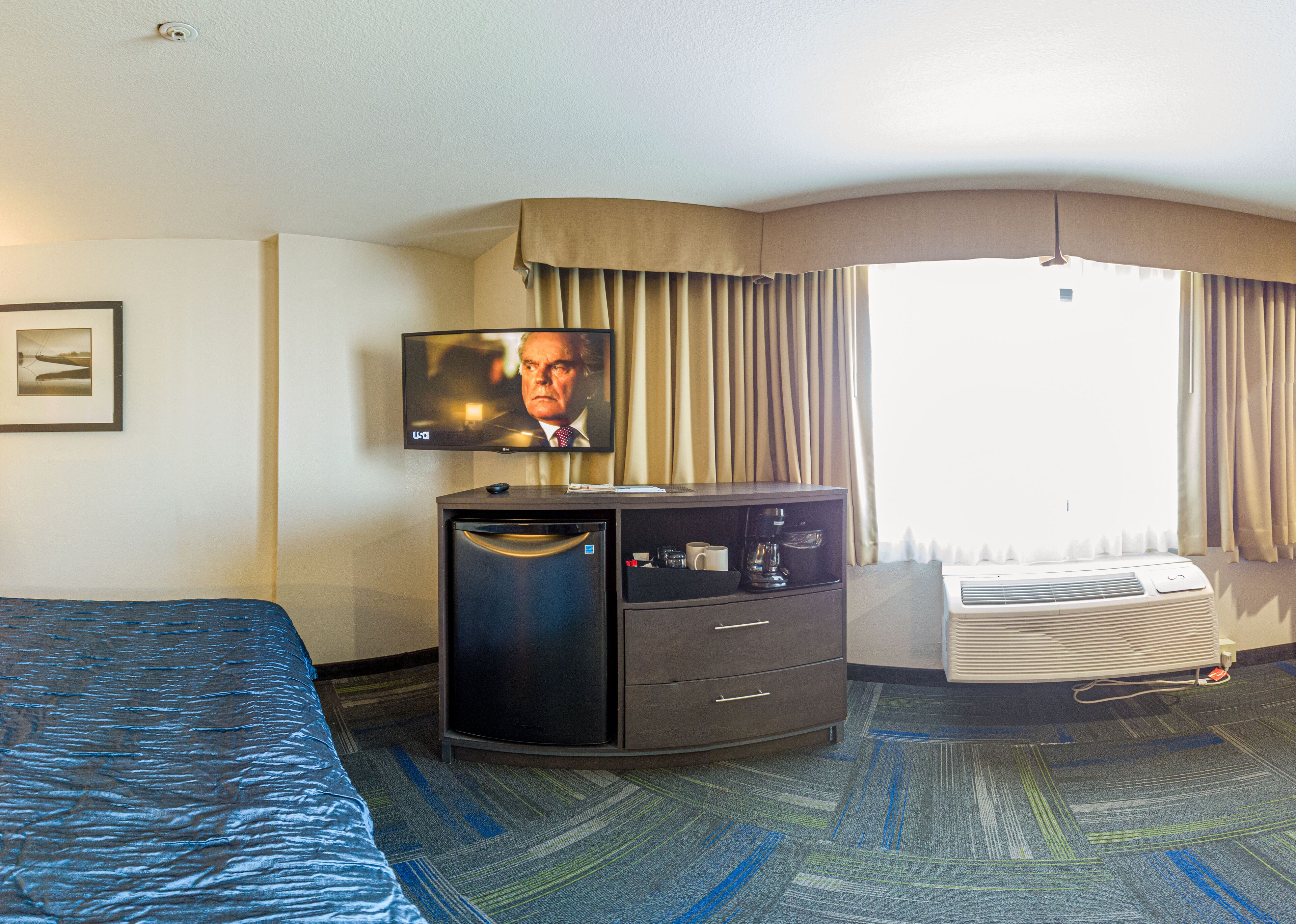A hotel room with a bed, a TV, a minibar, a desk, a window with curtains, and a carpeted floor.