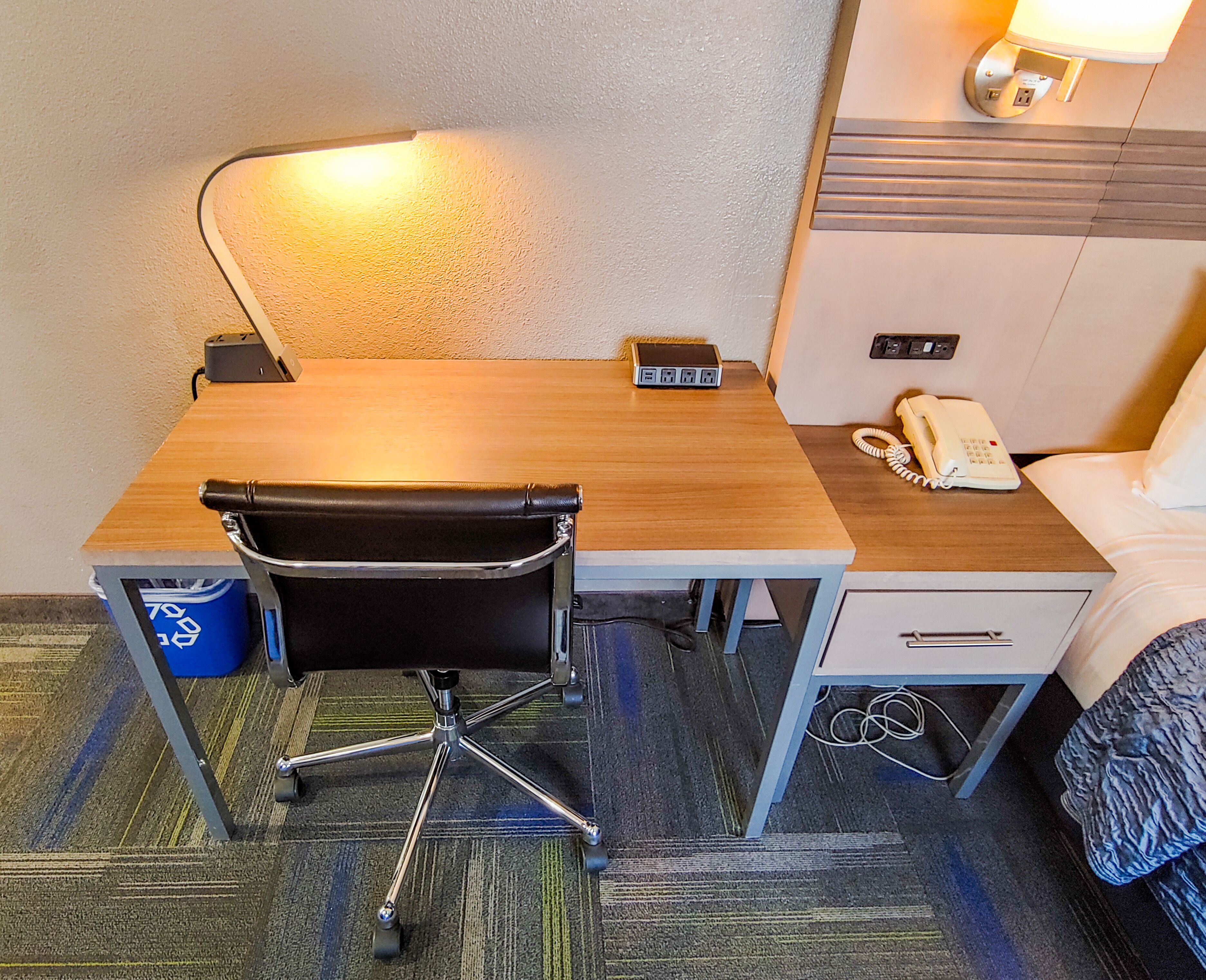 Une chambre d’hôtel avec un bureau en bois, une chaise de bureau noire, un téléphone, une table de chevet et une lampe.