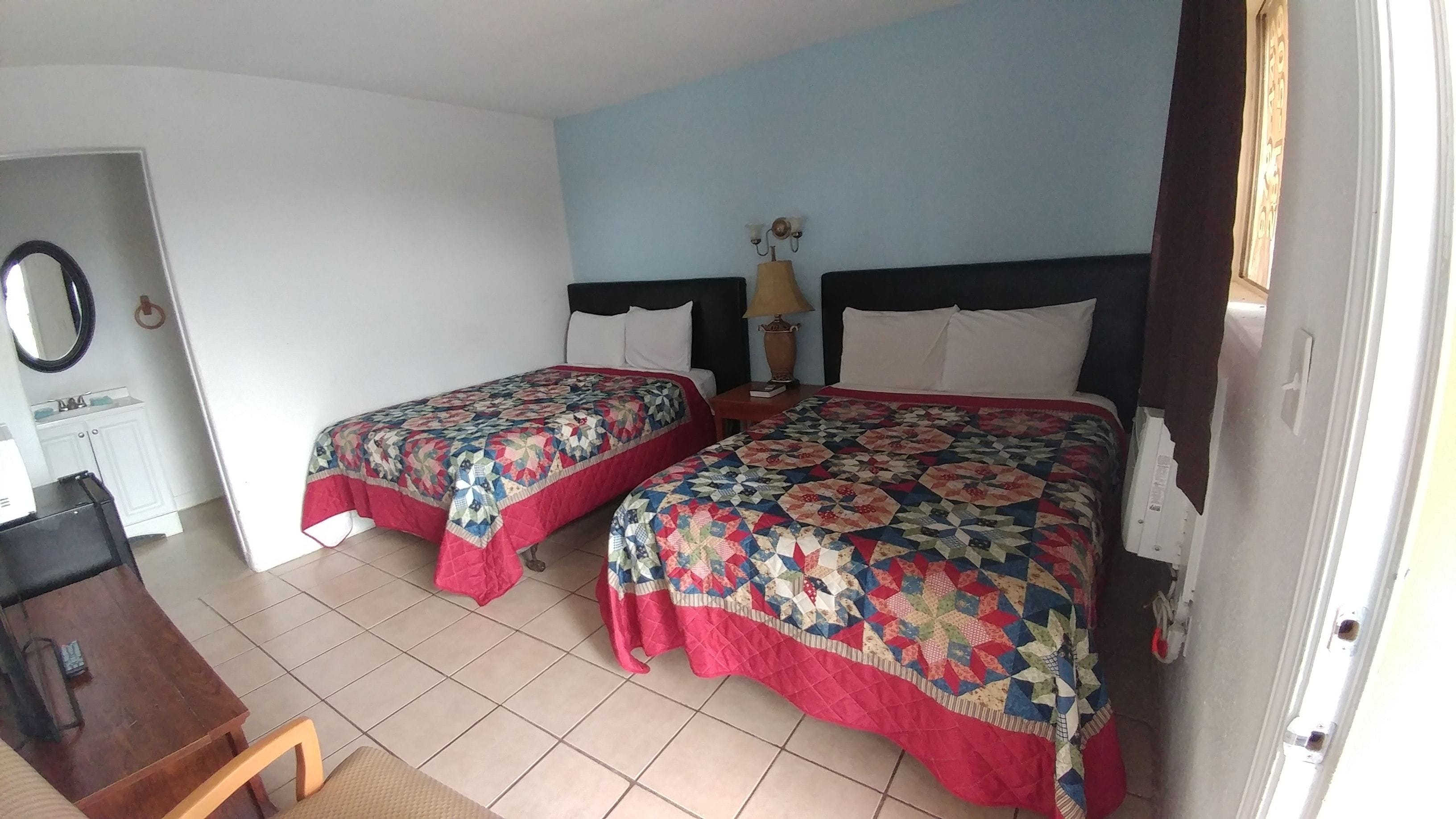 A hotel room with two beds, a desk, a chair, a lamp, and a window with curtains.