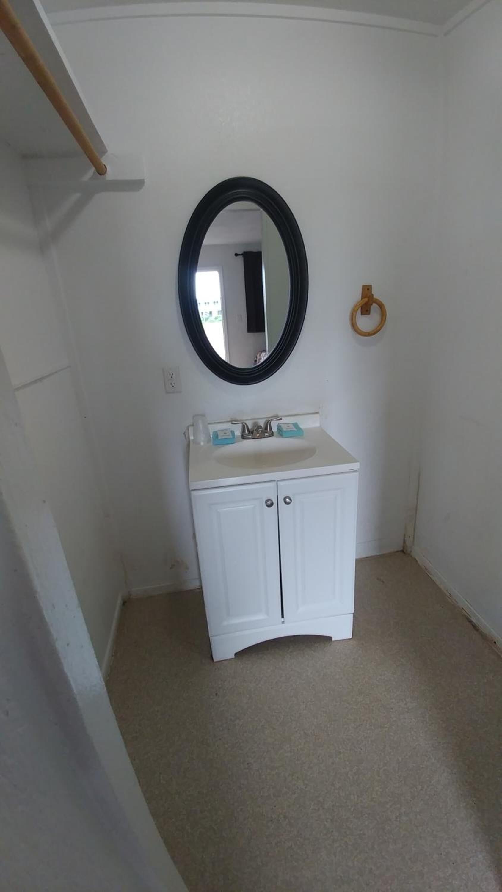Een kleine, witte badkamer met een ronde spiegel, een wit wastafeltje en een handdoekhouder aan de muur.