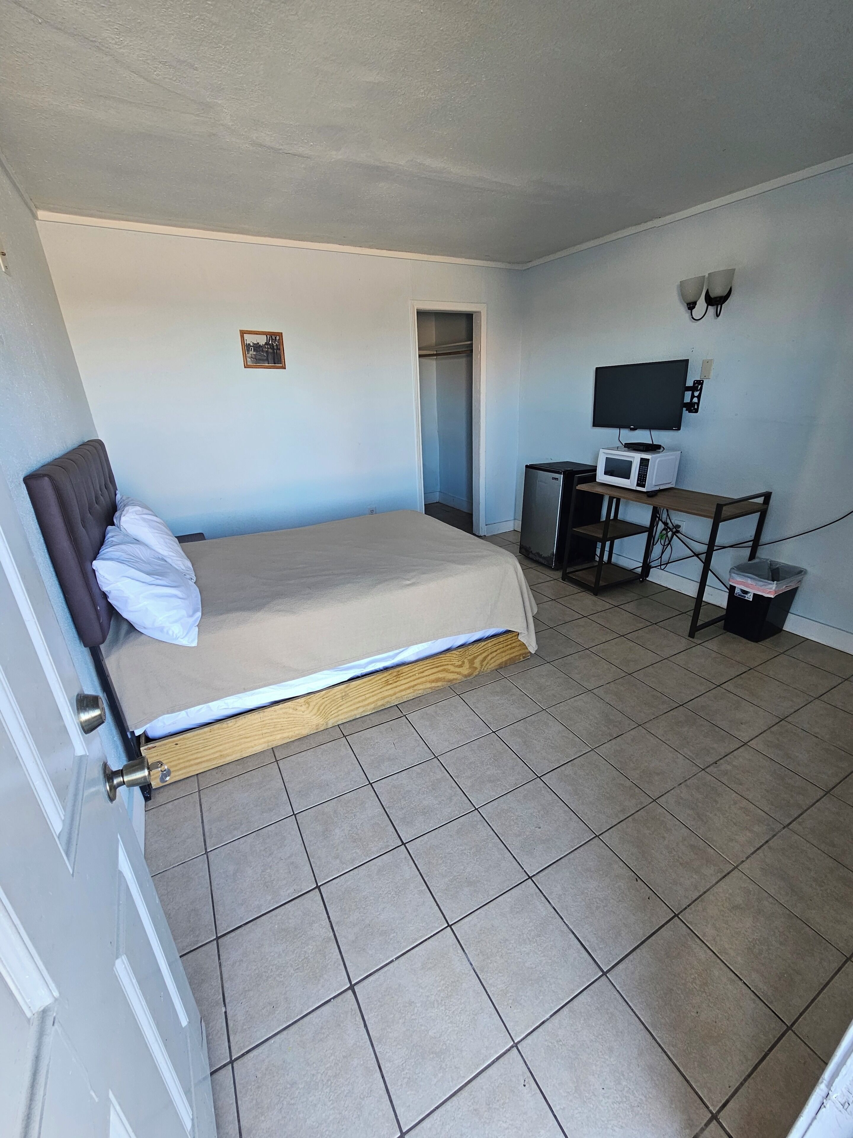 Ein Schlafzimmer mit Bett, Schreibtisch mit Computer und Mikrowelle, Kleiderschrank und kleinem, an der Wand befestigtem Fernseher.