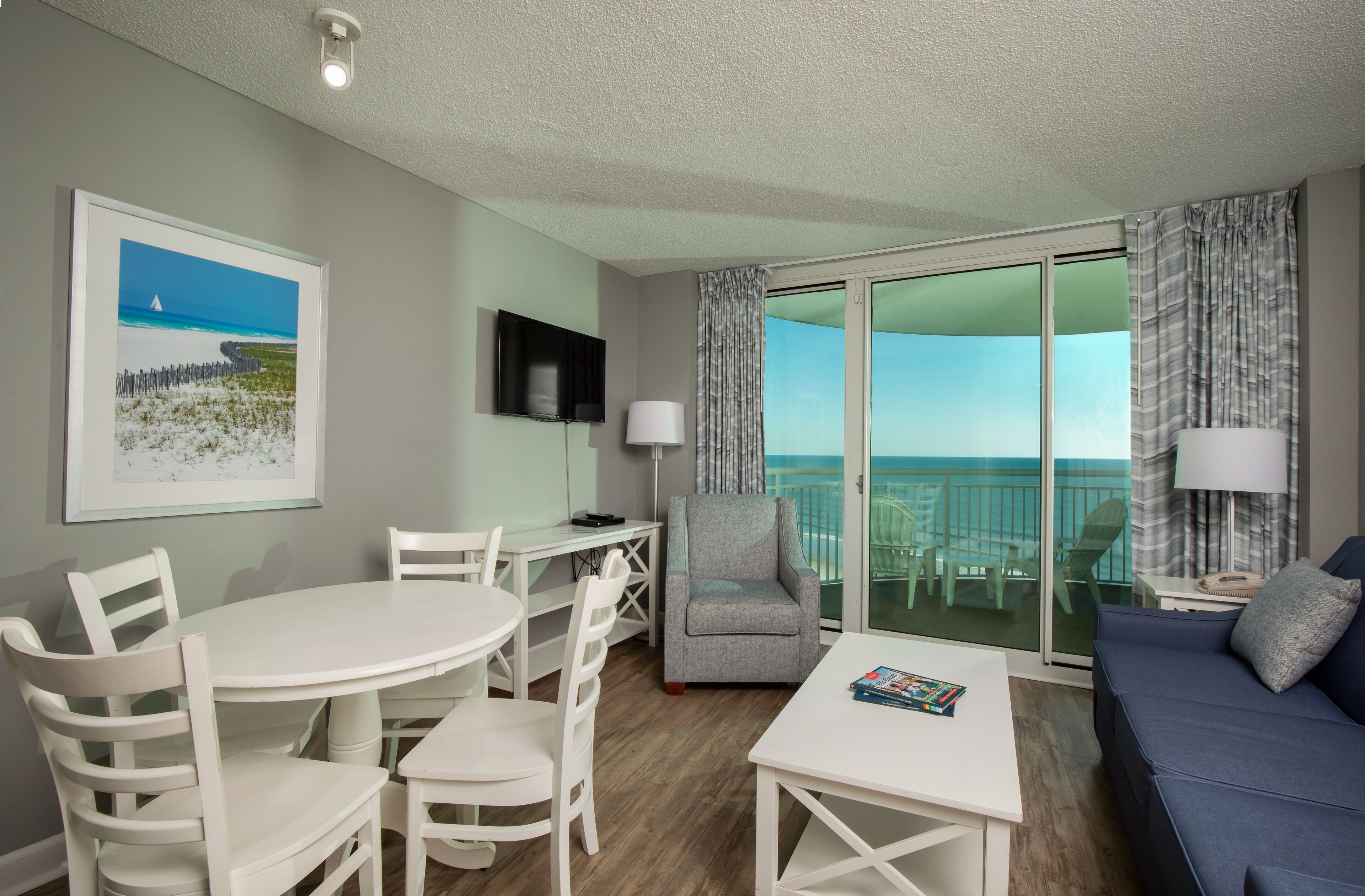 Habitación de hotel moderna con balcón, muebles blancos, televisor de pantalla plana y un cuadro de una playa.